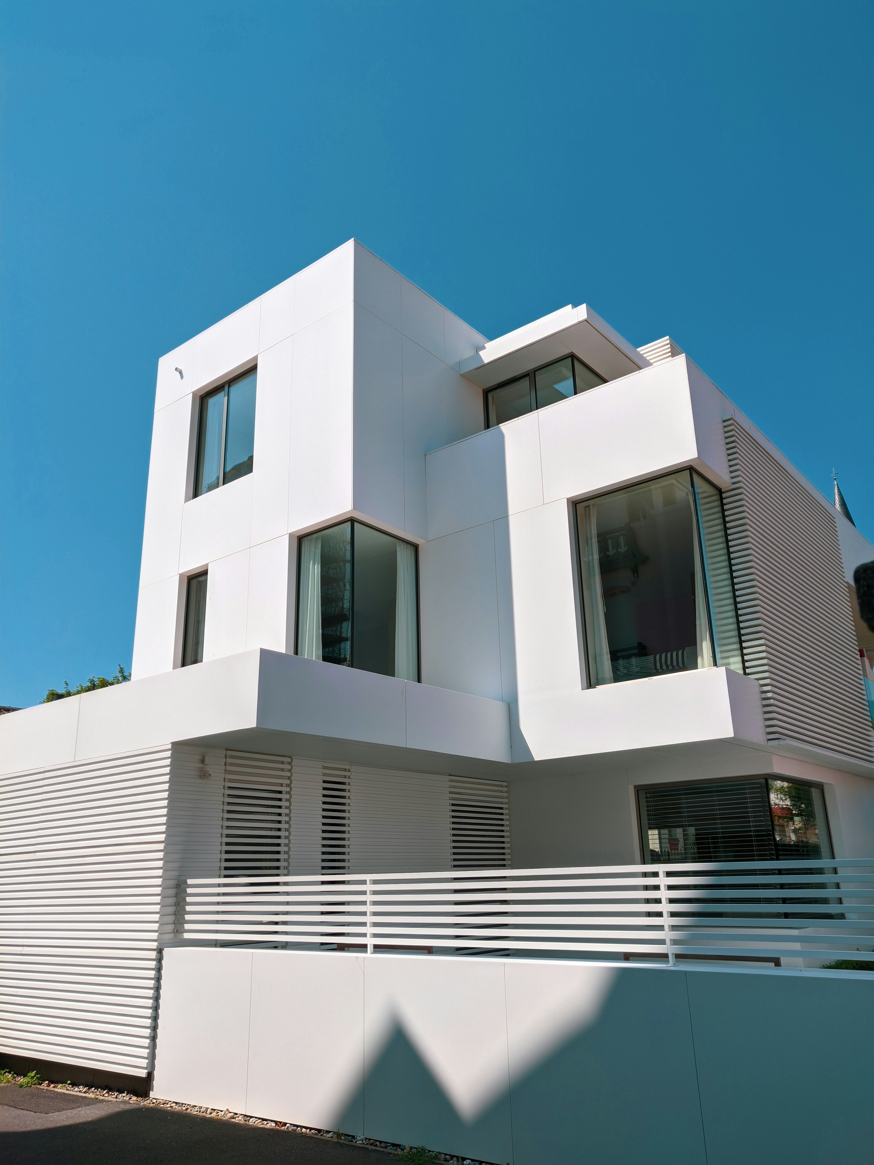 Sunlight and shadow across a white modern house facade, with projecting volumes, corner windows and horizontal slats. Photograph by Felix.